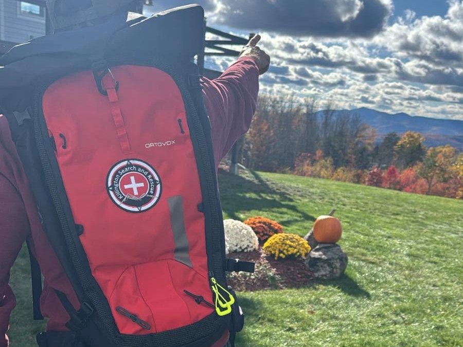 A KSAR member in a red Killington Search and Rescue backpack points toward the fall foliage and mountains