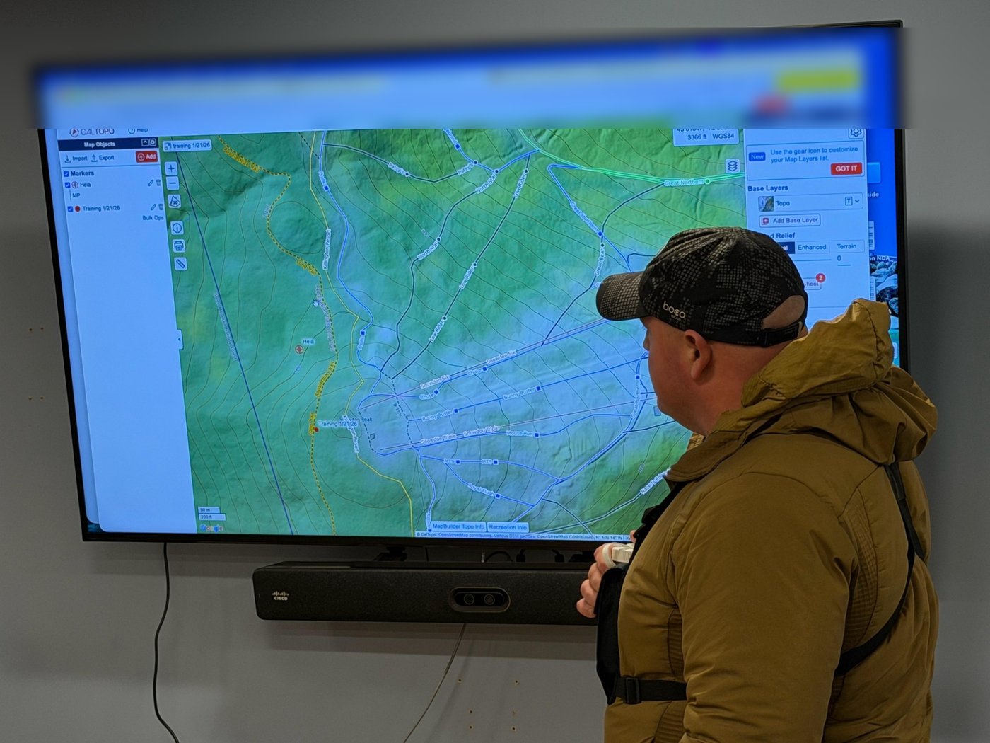 A KSAR member in a tan jacket and cap studies a CalTopo map of trails and ski runs near Killington Peak on a wall screen