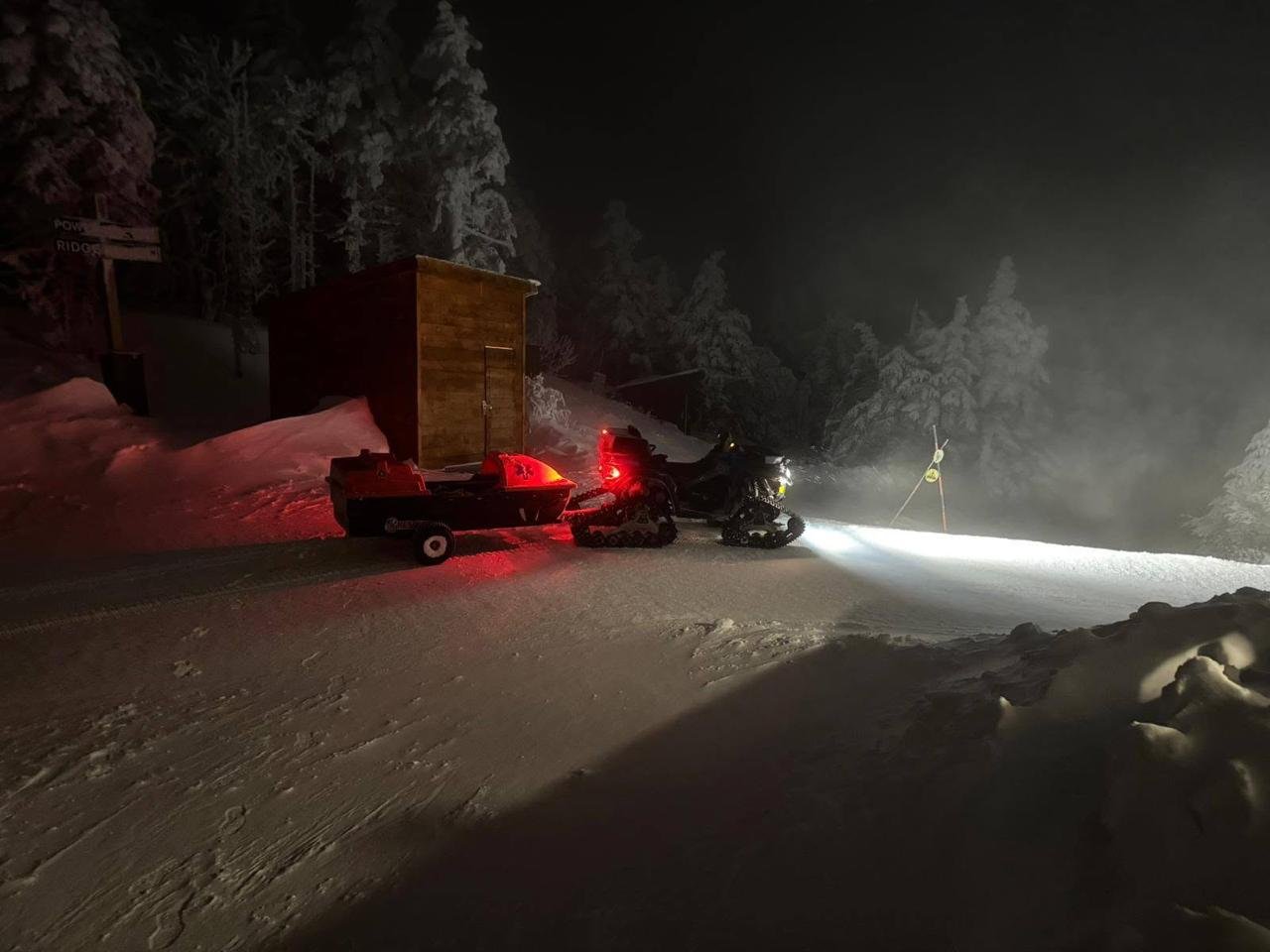 A KSAR snowmobile with a red rescue sled trailer parked beside a wooden shed on a snowy mountain road at night