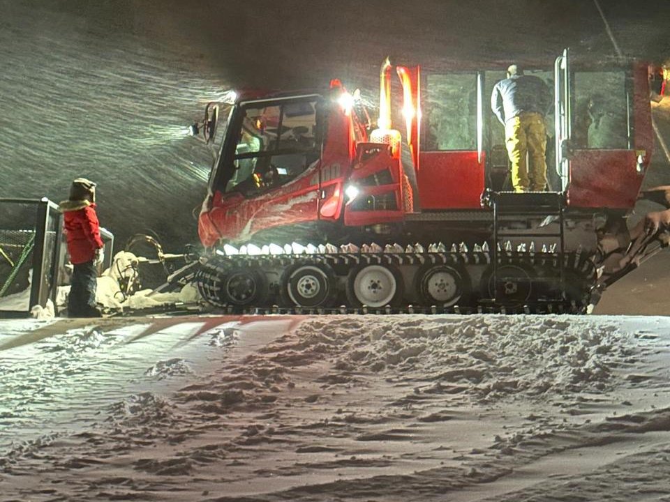 A red snowcat with its lights on works in blowing snow at night while a teammate in a red coat looks on