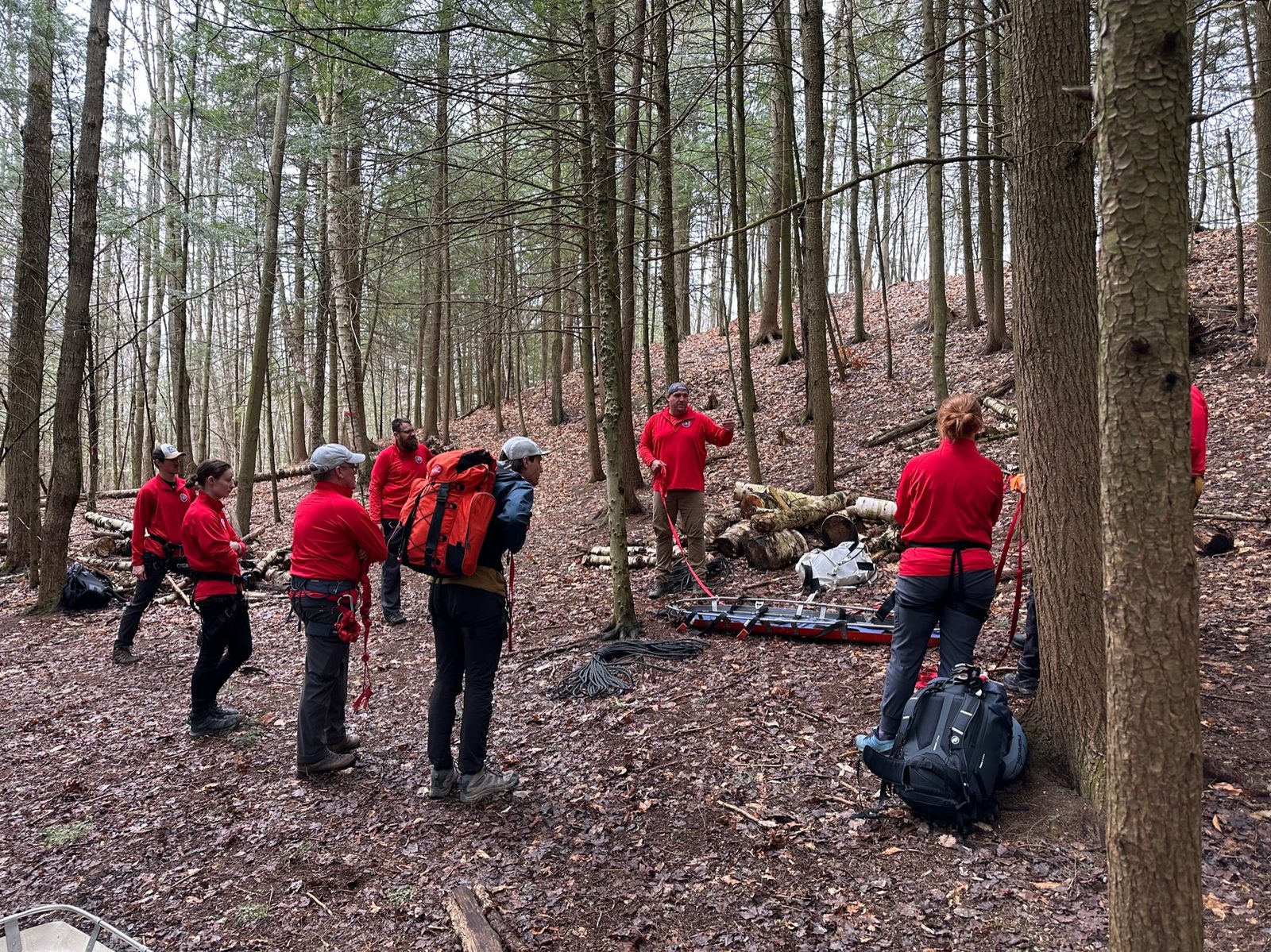 KSAR team members in red gather in the woods around a rescue litter during a training briefing